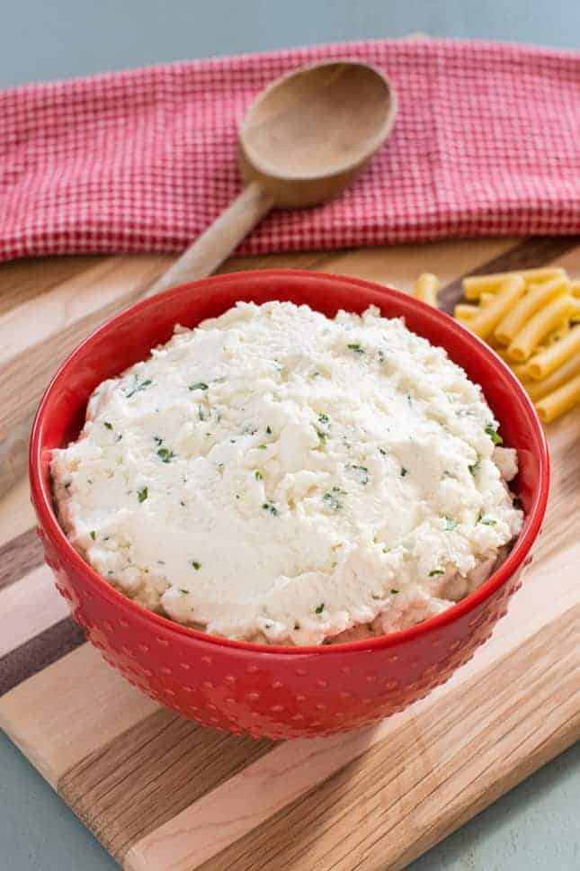 ricotta cheese mixture in a red bowl with a wooden spoon and some uncooked ziti noodles in the background
