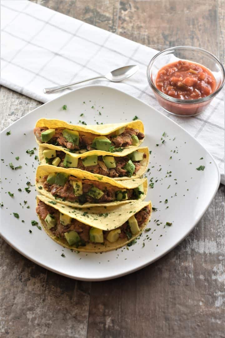 4 pinto bean tacos on a white plate topped with avocado and garnished with cilantro with salsa in the background