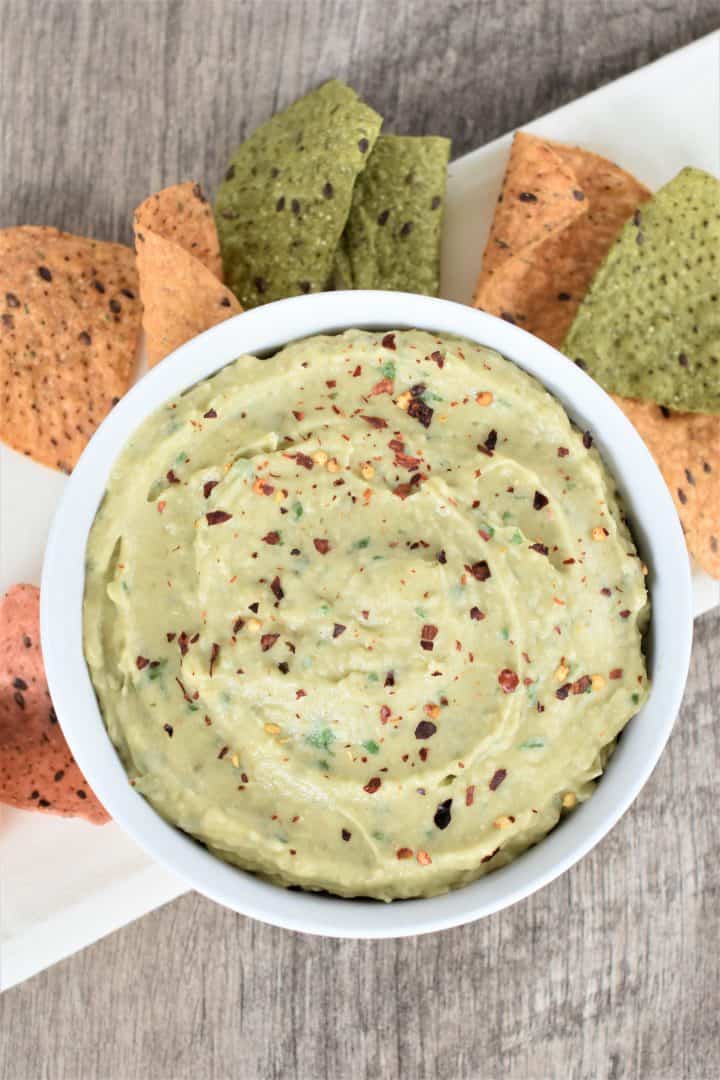 Overhead view of cannellini bean hummus with avocado in a white bowl with multicolored tortilla chips behind it