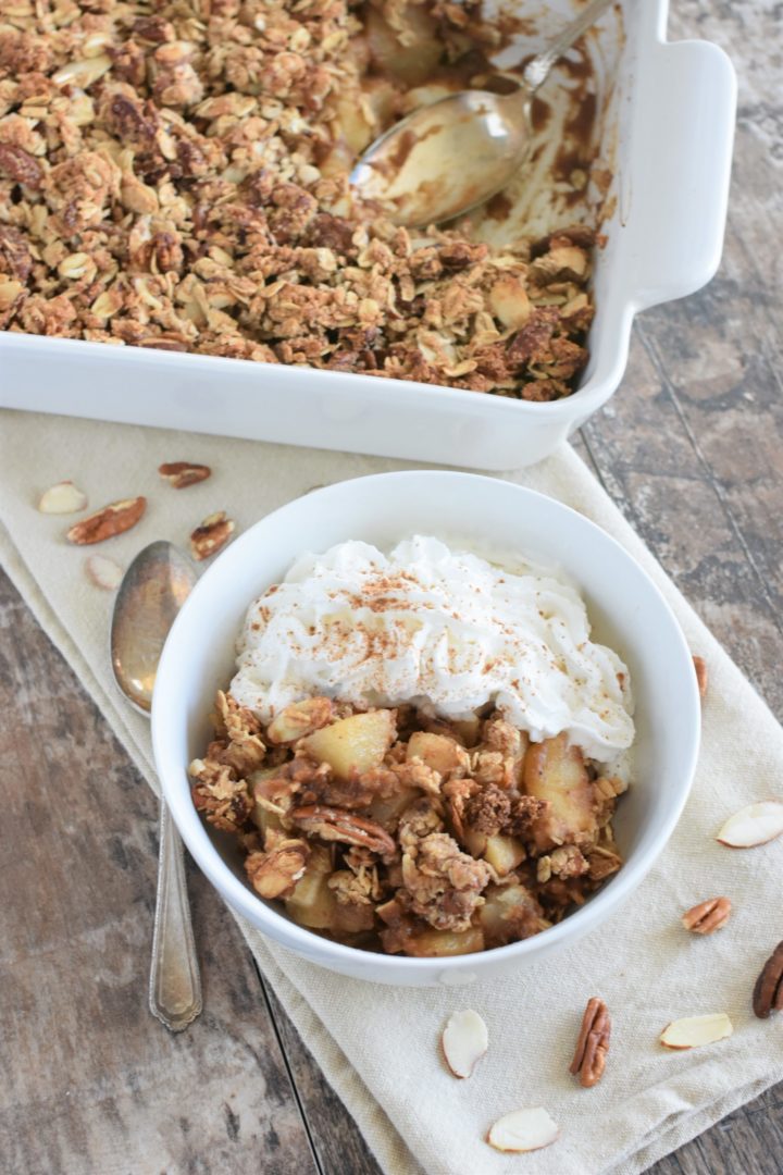 pear crisp in white bowl with coconut whipped topping and a spoon next to it with the rest in the baking dish in the background