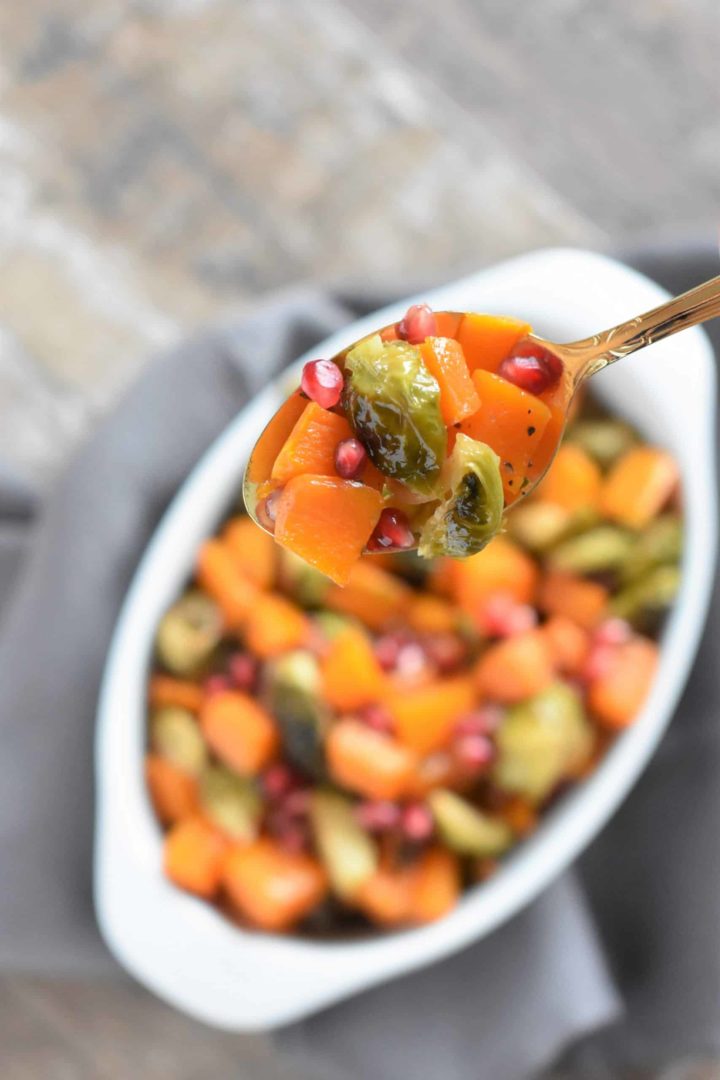 Spoonful of butternut squash and Brussels sprouts with pomegranate hovering over the rest in white serving dish