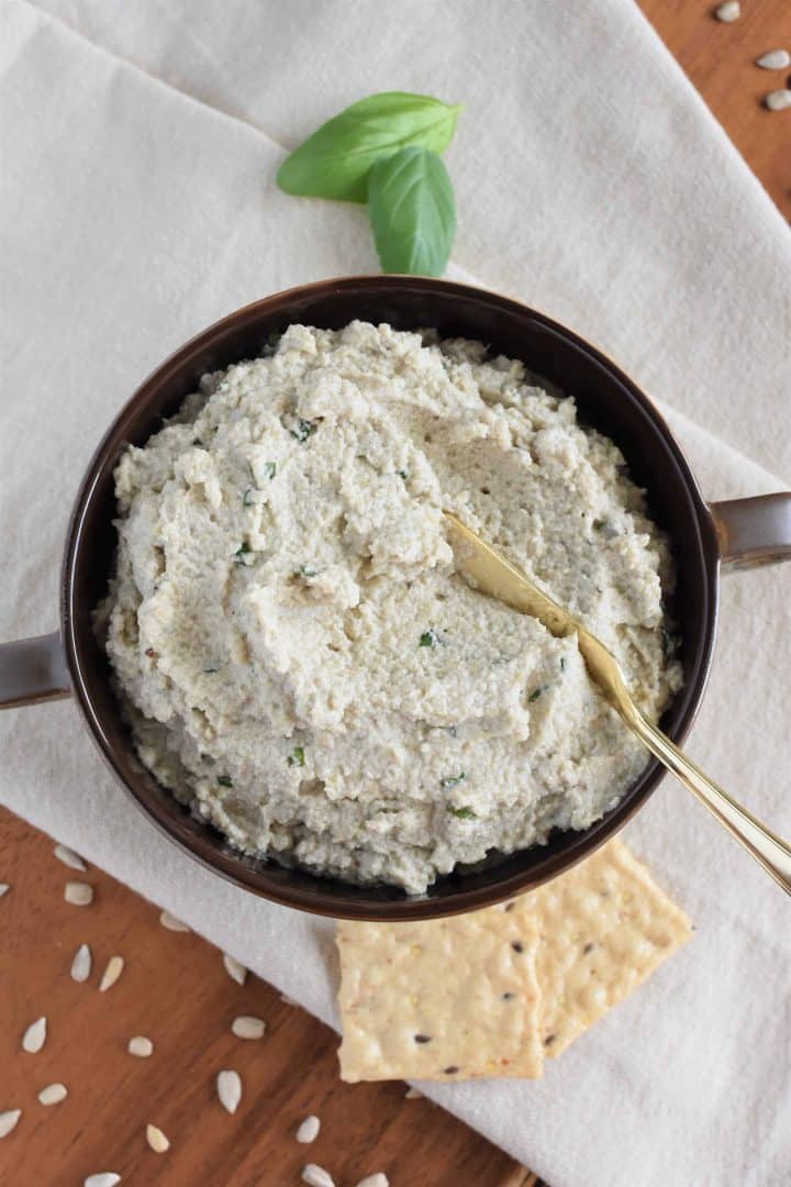 overhead of sunflower seed ricotta with spreading knife in the bowl and crackers, basil and sunflower seeds surrounding it