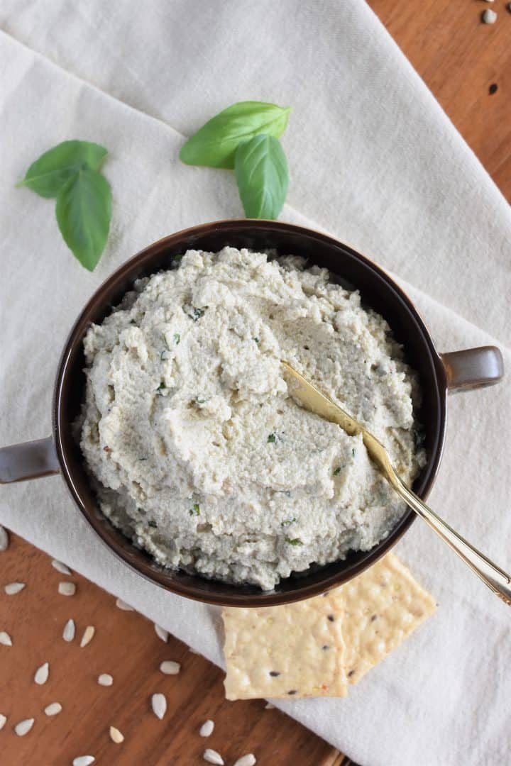 overhead of vegan ricotta with spreading knife in the bowl and basil, crackers and sunflower seeds surrounding it