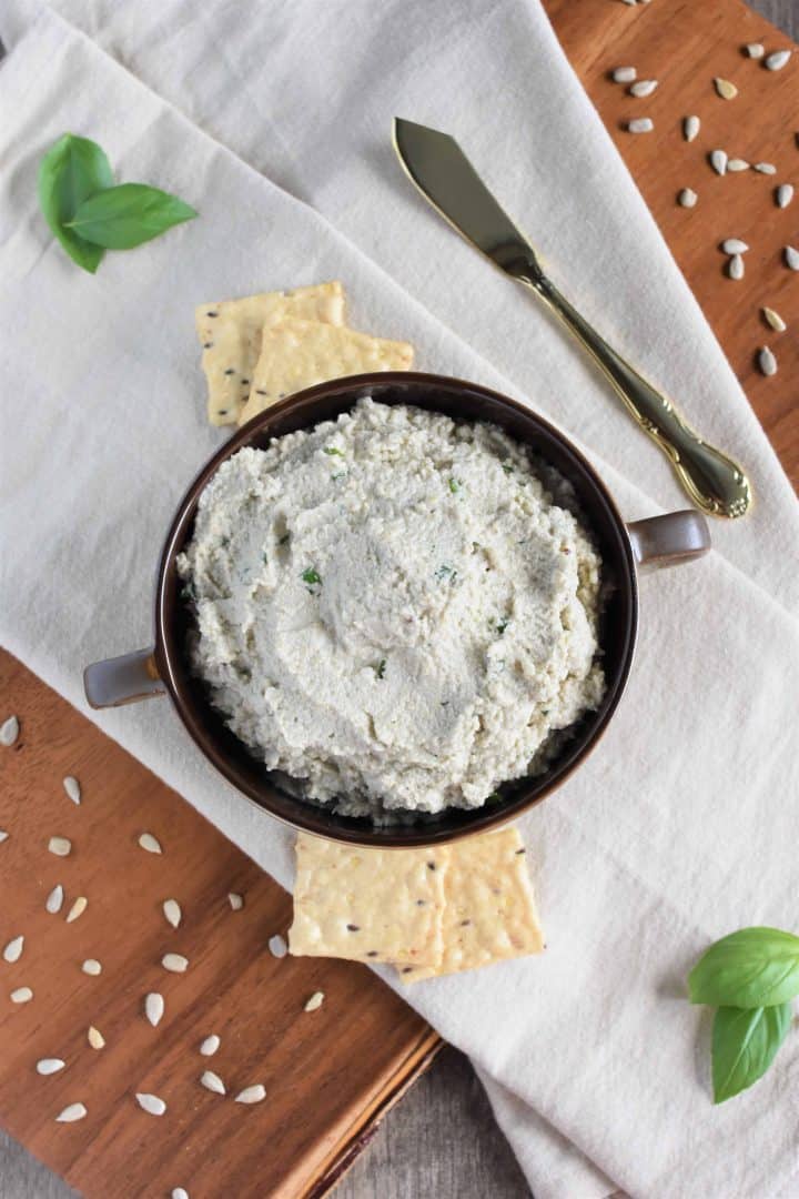 overhead of sunflower seed ricotta with crackers, basil and sunflower seeds surrounding it and knife next to it