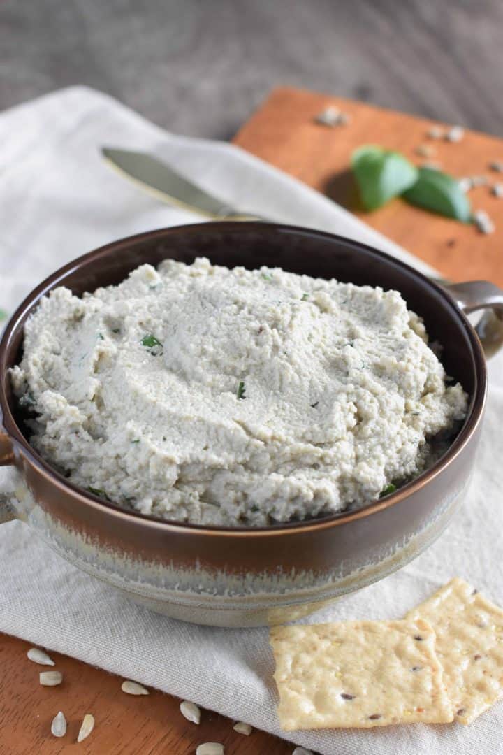 front view of sunflower seed ricotta with knife and basil in background and crackers in front and sunflower seeds sprinkled around