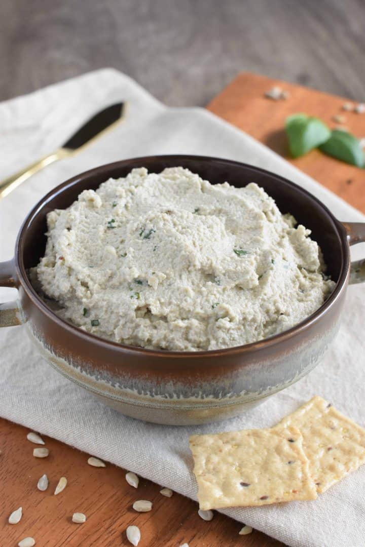 front view of sunflower seed ricotta with crackers, basil and sunflower seeds surrounding it and knife in background