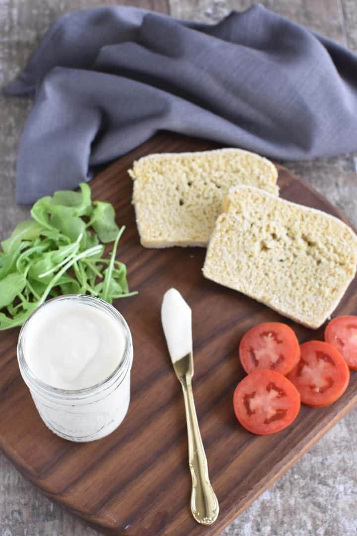 view of mayo in mason jar with spreading knife, arugula, tomatoes and bread all on a wooden board