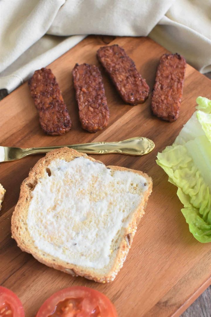 mayo spread on one piece of toast with tomatoes, tempeh bacon and lettuce next to it all on a wooden board