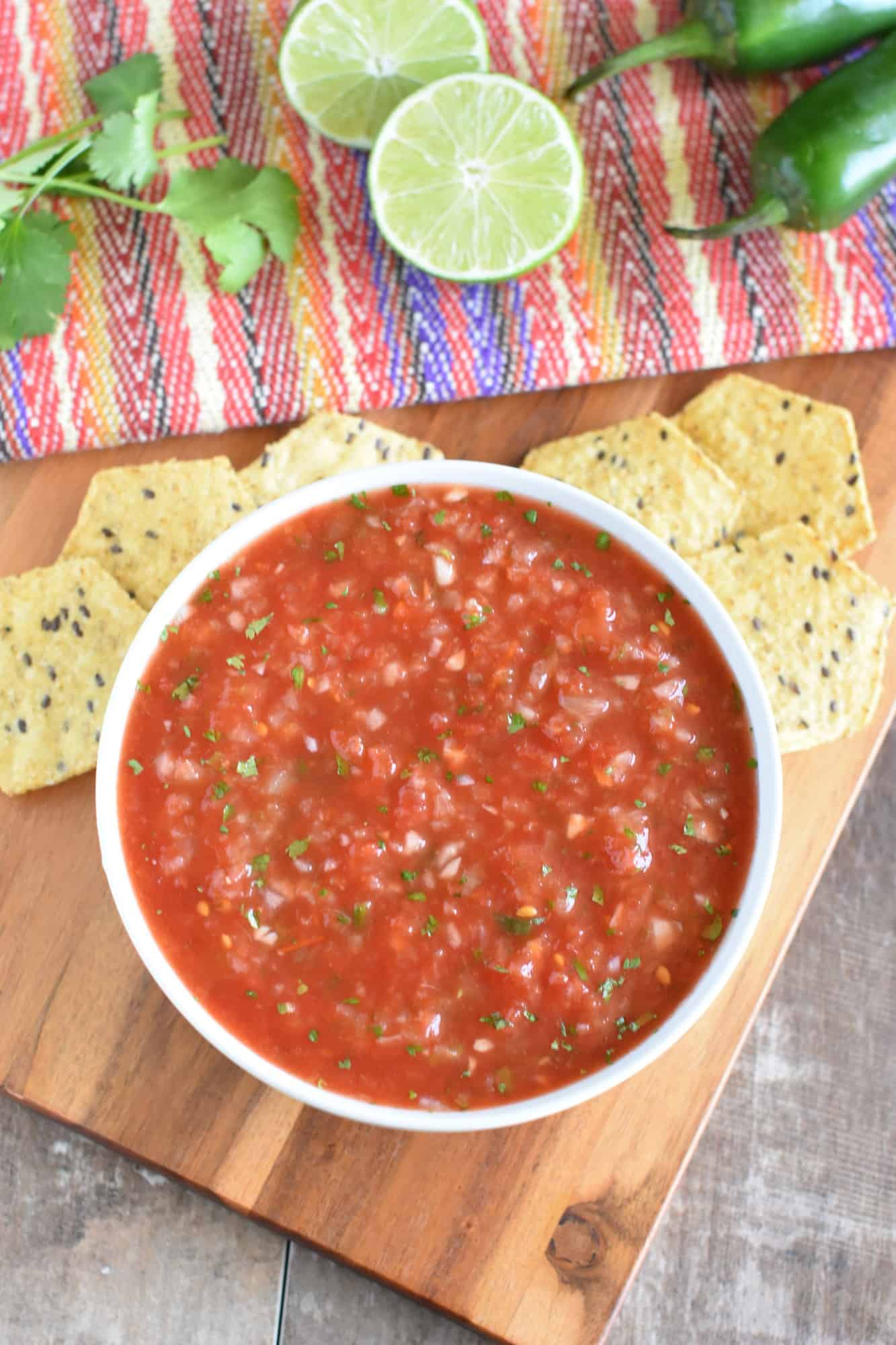 overhead of salsa in white bowl with chips on a wooden board and cilantro, lime and jalapeno behind the chips