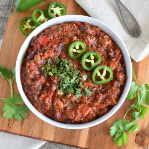 overhead of chili in a white bowl on a wooden board with cilantro and jalapenos