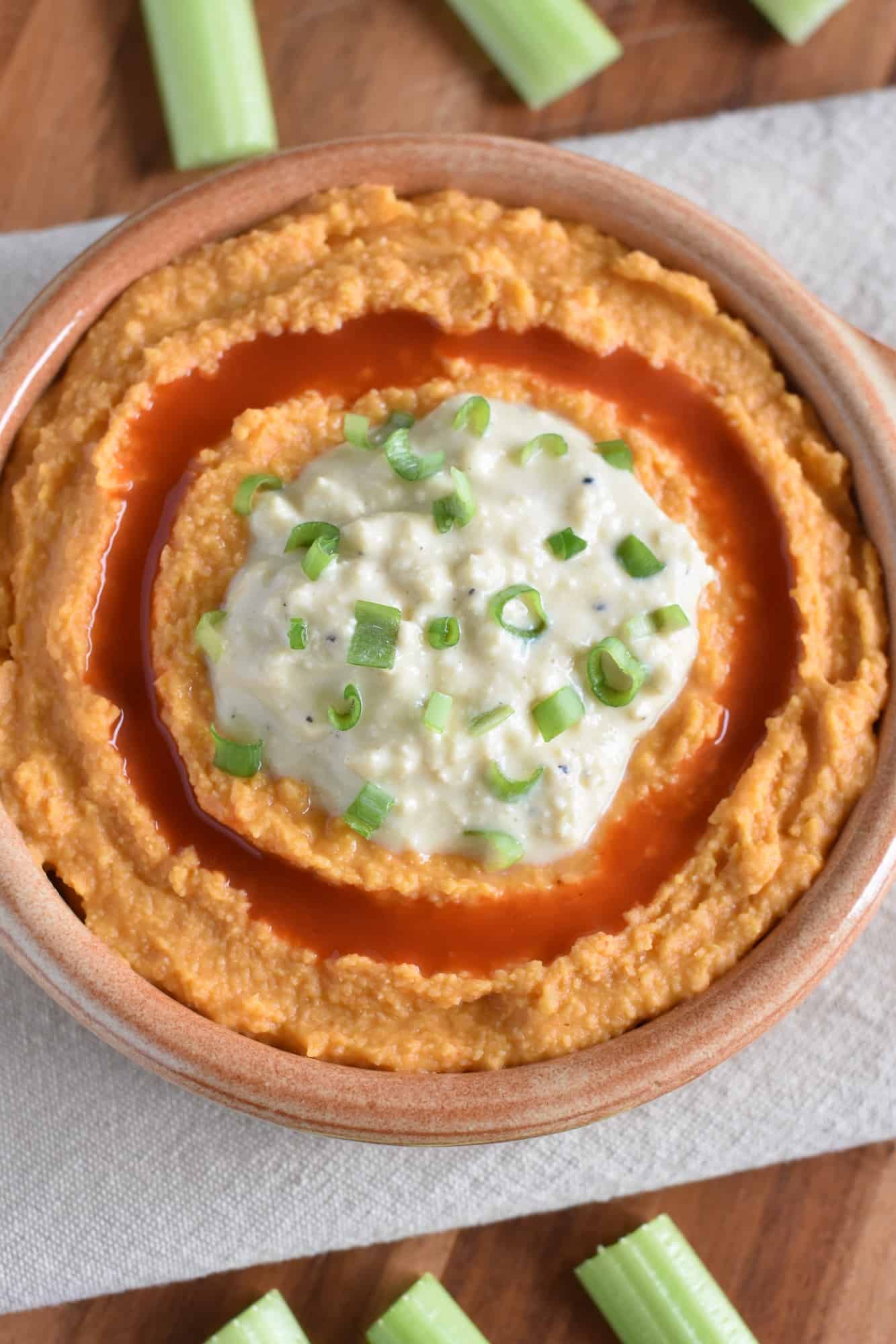 hummus in serving bowl on kitchen towel on wooden board with celery sticks around it