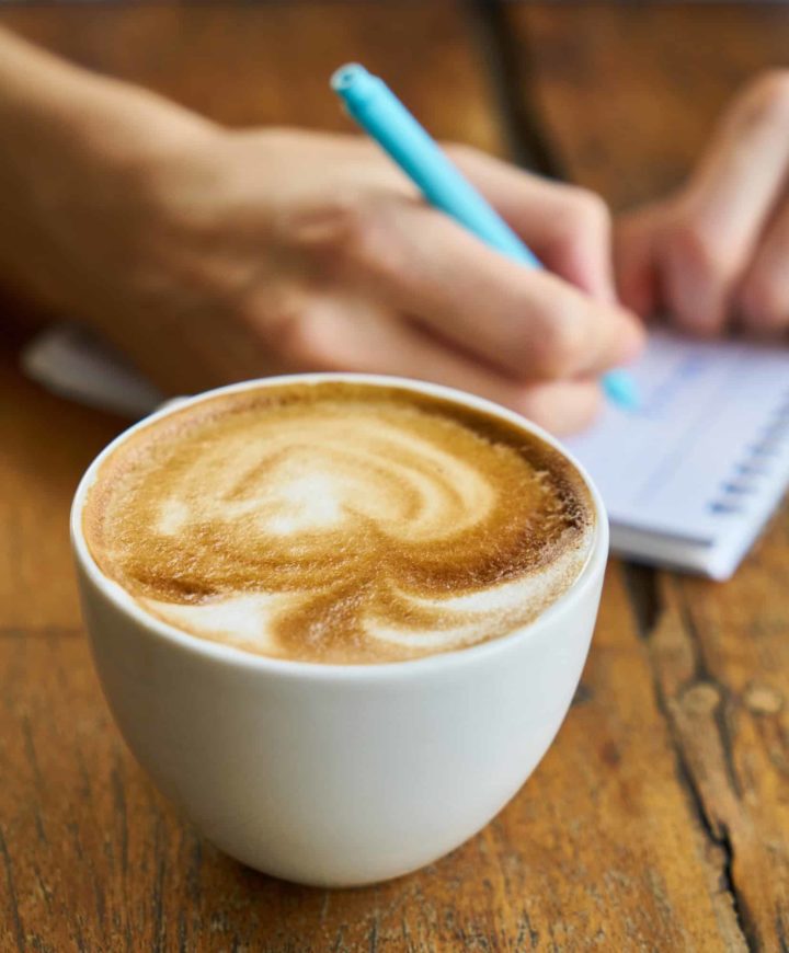 person writing in a small notebook with a latte next to the notebook