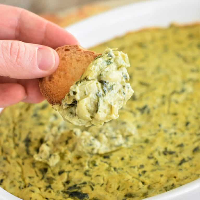 close-up of hand holding bread with dip on it over the casserole of the rest of the dip