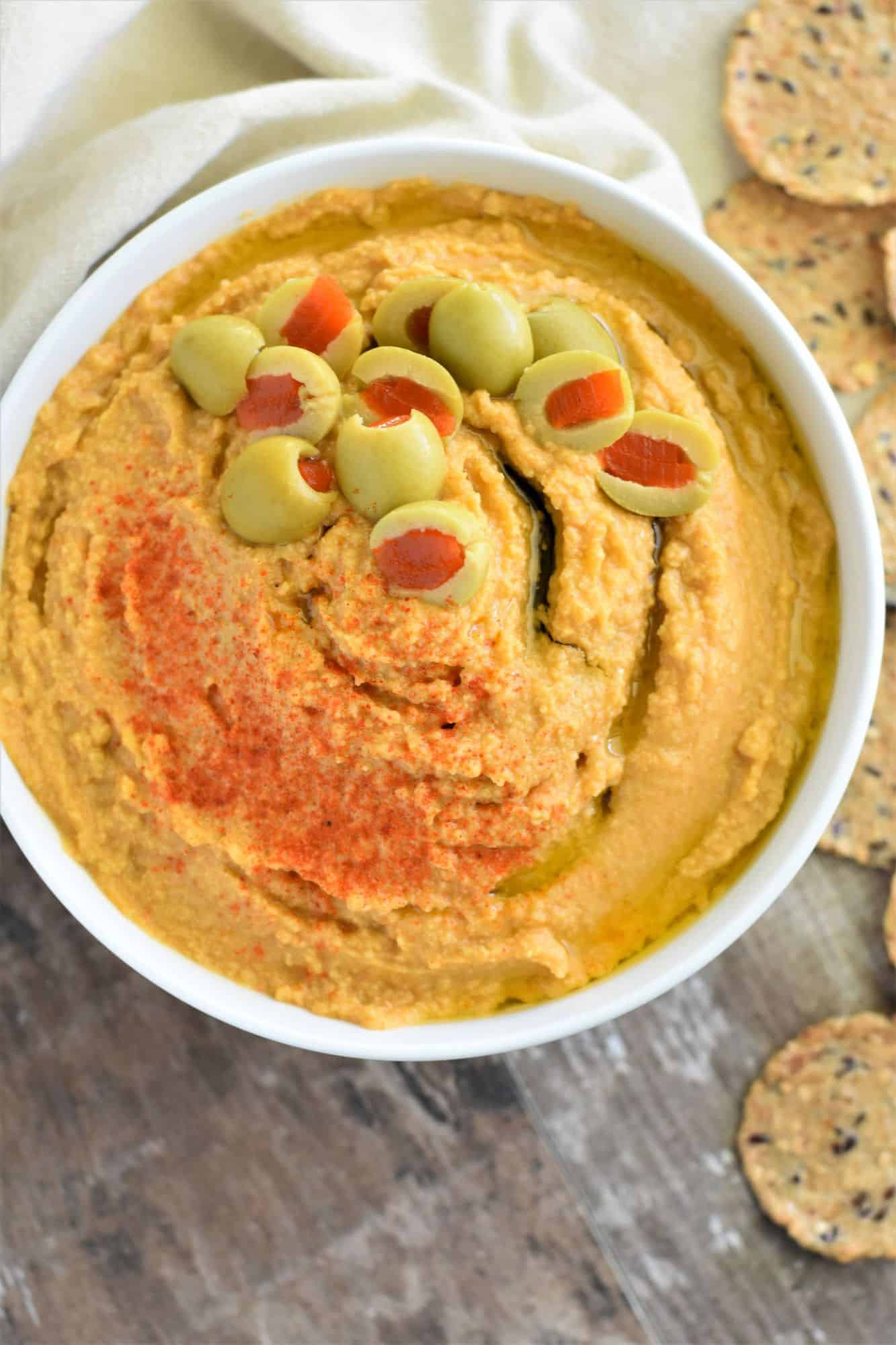 overhead of green olive hummus in white bowl with crackers around it