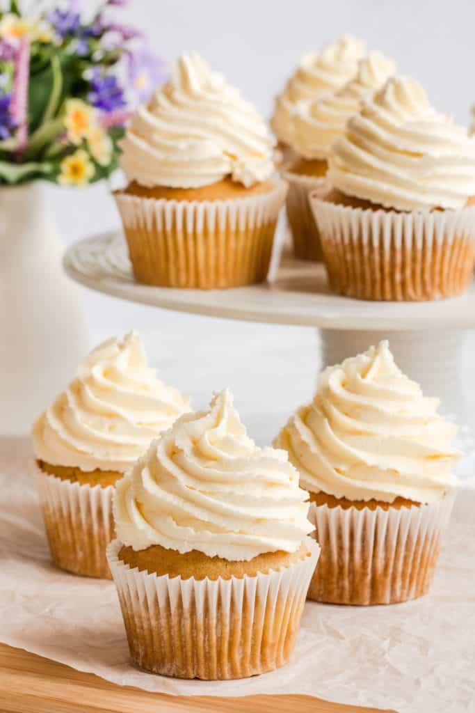 3 frosted cupcakes on a piece of parchment with more on a raised cake plate behind them