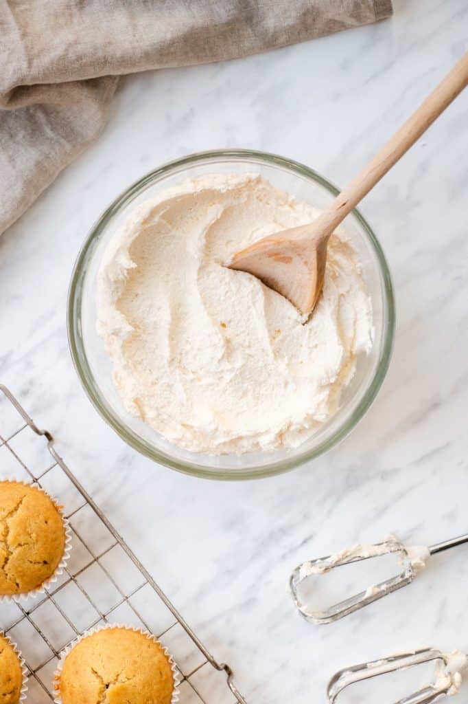 overhead of frosting in mixing bowl with wooden spoon in it and some unfrosted cupcakes waiting to be iced