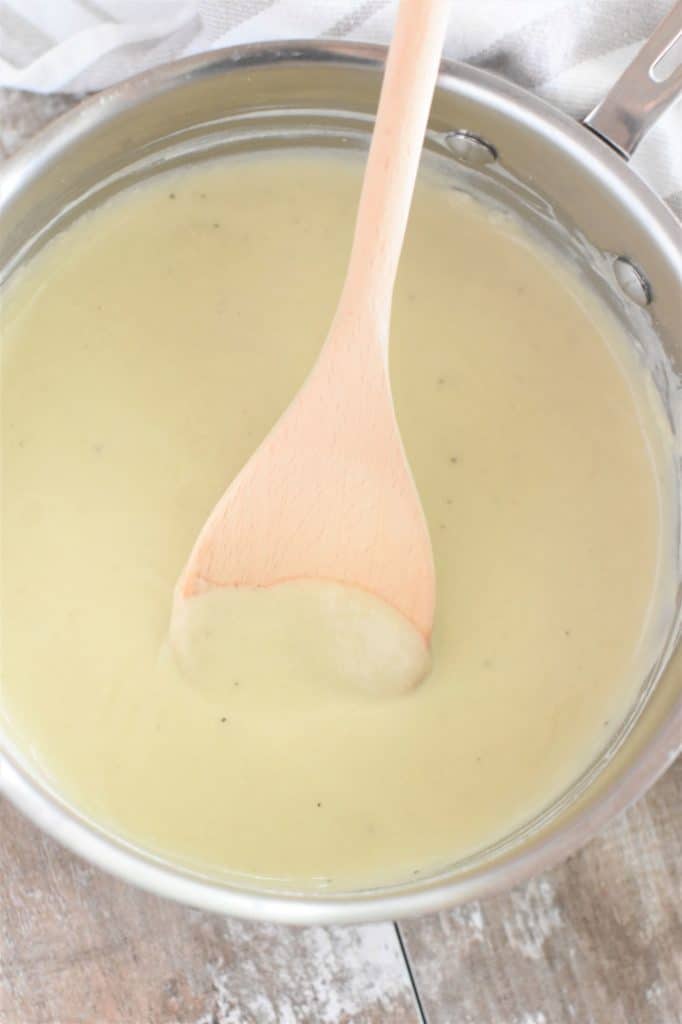 overhead of saucepan of vegan bechamel with a wooden spoon in it and a kitchen towel behind it