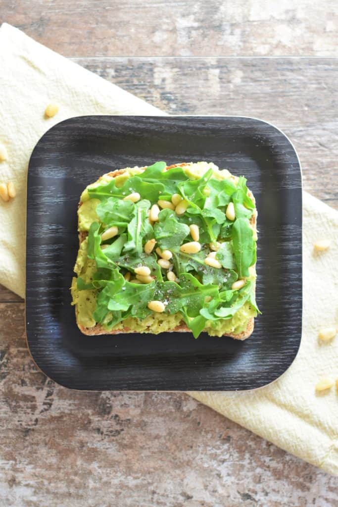 overhead of slice of avocado toast with arugula and pine nuts