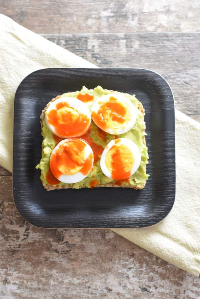 overhead of slice of avocado toast with boiled eggs and hot sauce