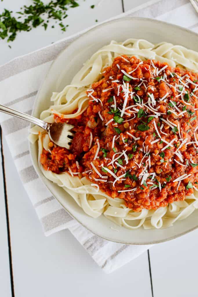 close-up overhead of pasta with sauce on a plate garnished with parsley and Parmesan and a fork going in from the left