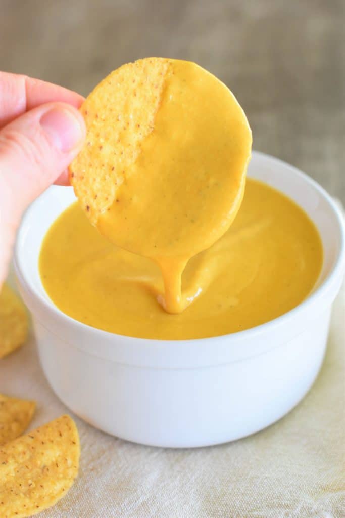 holding a tortilla chip with cheese sauce dripping off of it over the bowl of the rest of it