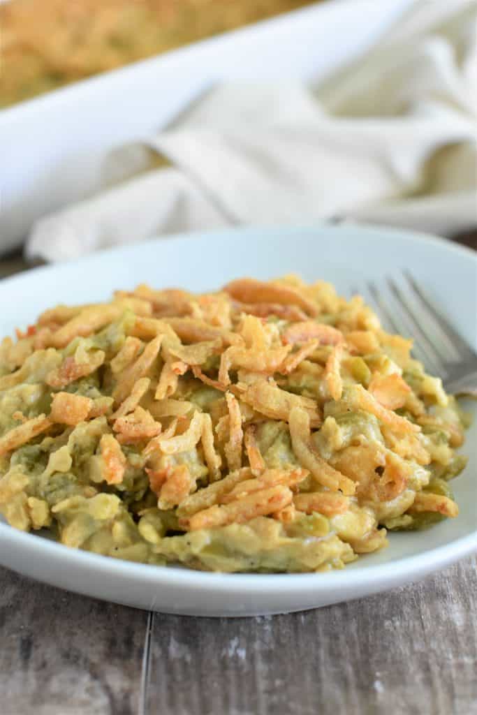 front view of serving of vegan green bean casserole on white plate with rest of it in baking dish blurred out behind it