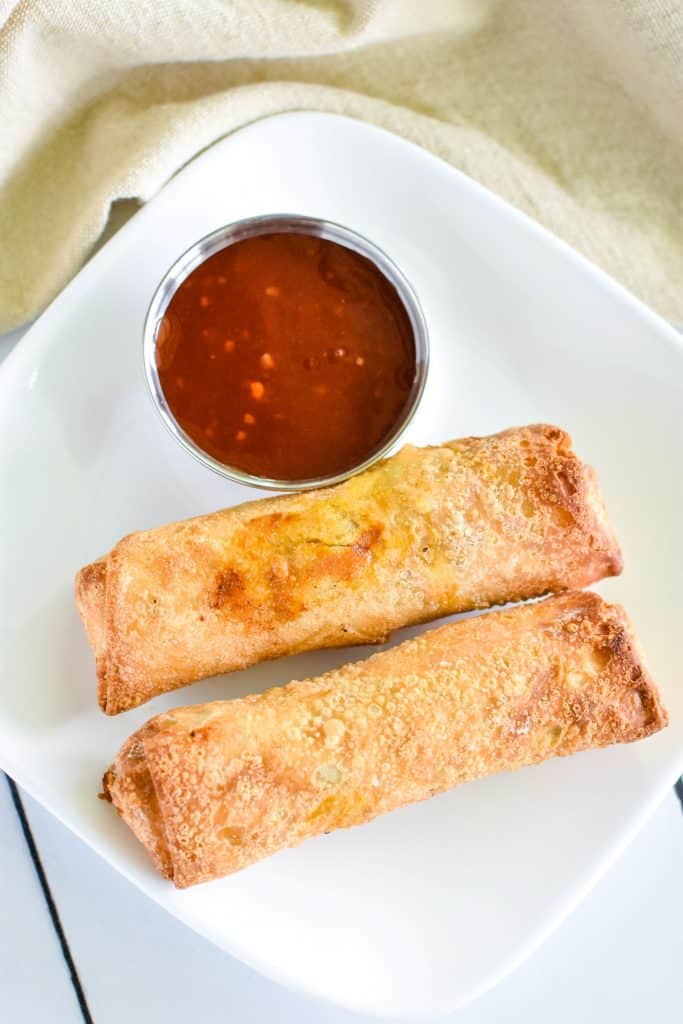 overhead of two egg rolls on a white plate with sweet chili sauce on the side.