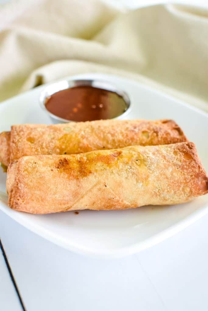 front view of two egg rolls on a white plate with side of sweet chili sauce.