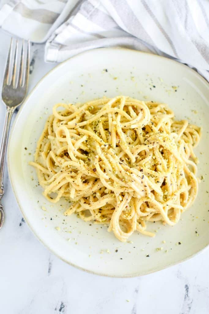 overhead of pasta on a white dish garnished with pepper and pumpkin seed Parmesan with a fork to the left of the dish