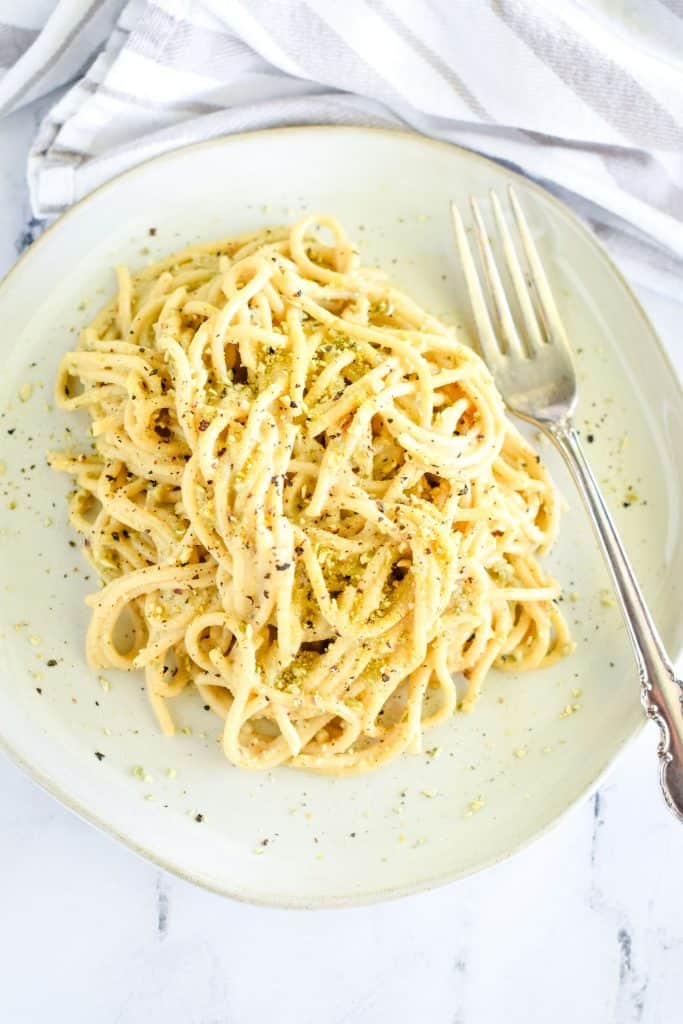 overhead of pasta on a white plate garnished with pepper and pumpkin seed Parmesan with fork on the plate on the right side