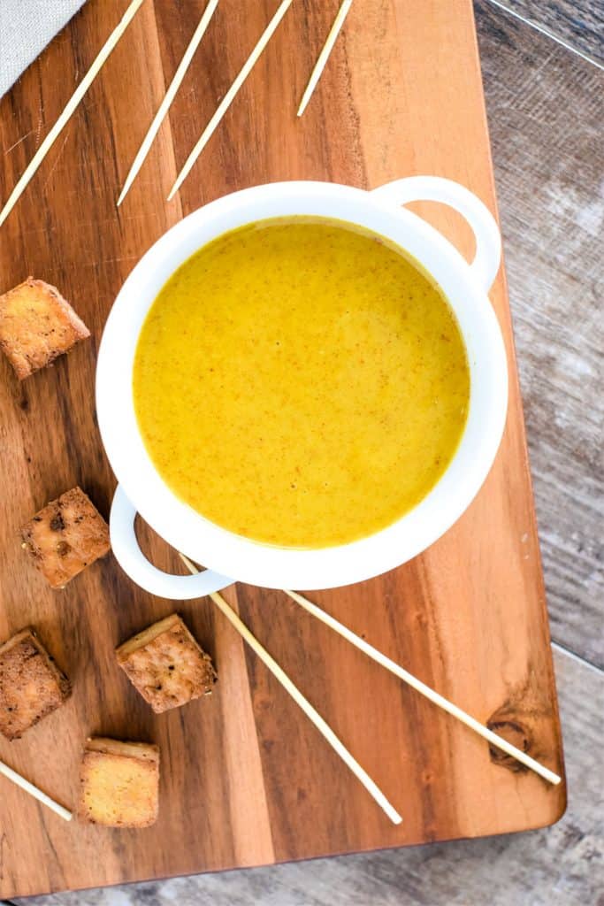 Honey mustard in white serving bowl with tofu pieces and skewers around it on wooden block