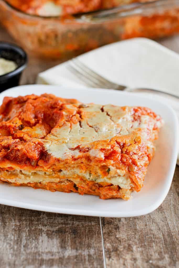 front view of slice of lasagna on a white plate with fork on a napkin, vegan Parmesan and the rest of the dish behind it