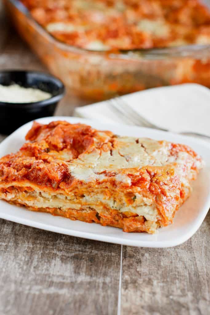 front view of slice of lasagna on white plate with vegan Parmesan, fork on a napkin and the rest of the food behind it