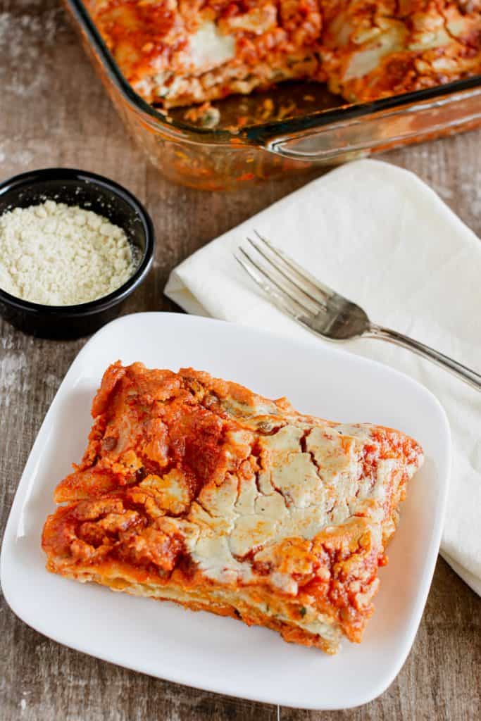 overhead of slice of lasagna on a white plate with vegan Parmesan, fork on a napkin and part of the rest in a baking dish in view behind it