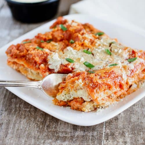 fork cutting into a piece of lasagna on a white plate topped with vegan Parmesan and fresh parsley