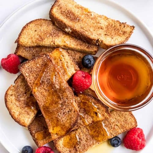 overhead of French toast sticks on a white plate topped with syrup and some berries and more syrup on the plate