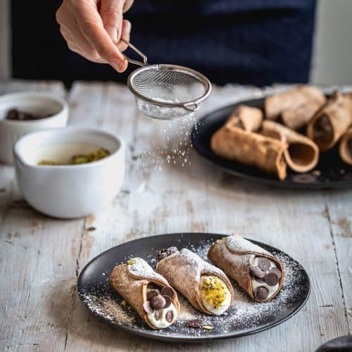 3 cannoli on black plate with powdered sugar being sifted on