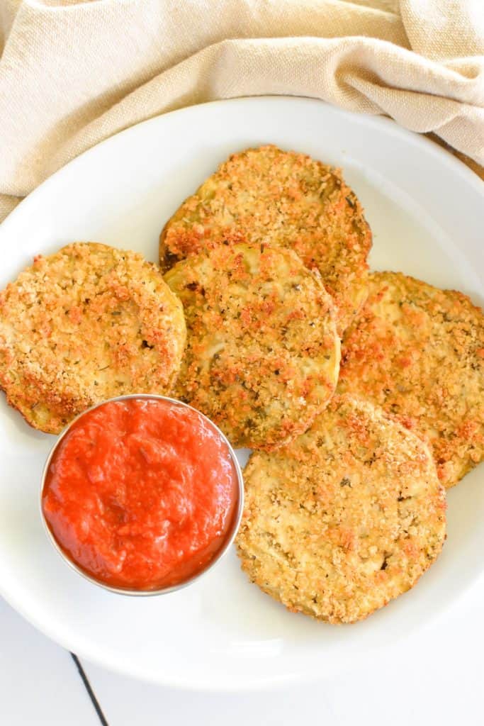 close-up overhead of eggplant slices on white plate with cup of sauce toward bottom of plate