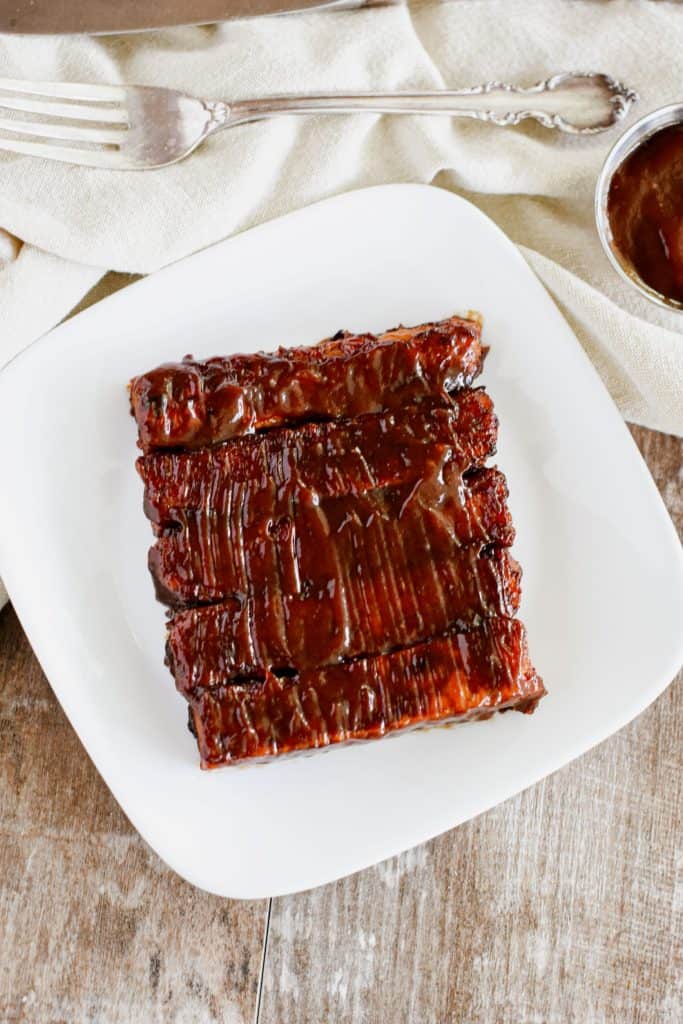 overhead of 5 tempeh ribs on a white plate with BBQ sauce slathered on top of them.