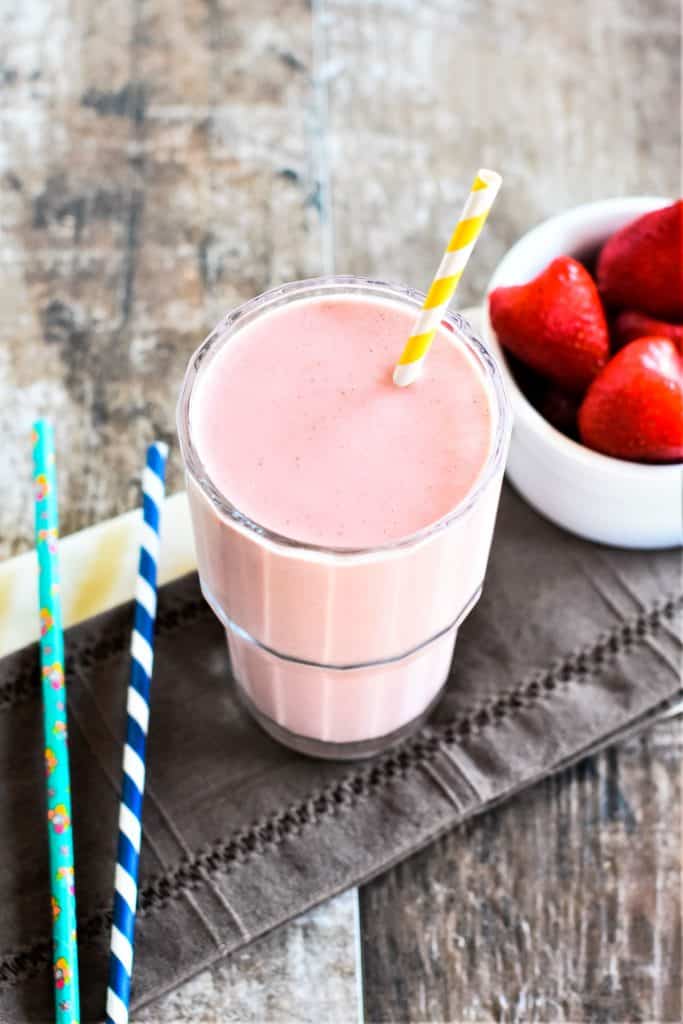 slight overhead of smoothie in a glass with a straw in it and straws and strawberries off to the sides