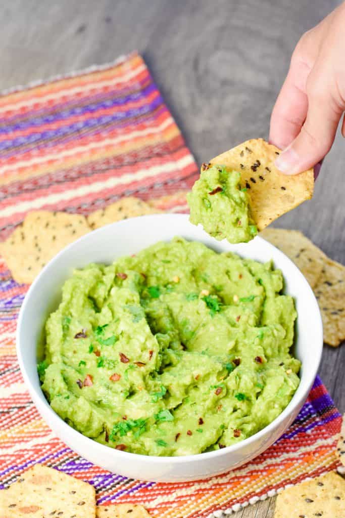 hand holding up a chip with guacamole dip on it over the bowl.