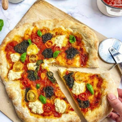 hand taking a slice of vegan pesto pizza from the rest on a pizza stone.