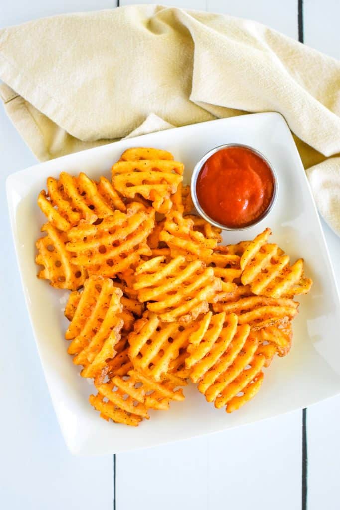 overhead of fries on a white plate with ketchup and tan napkin behind.