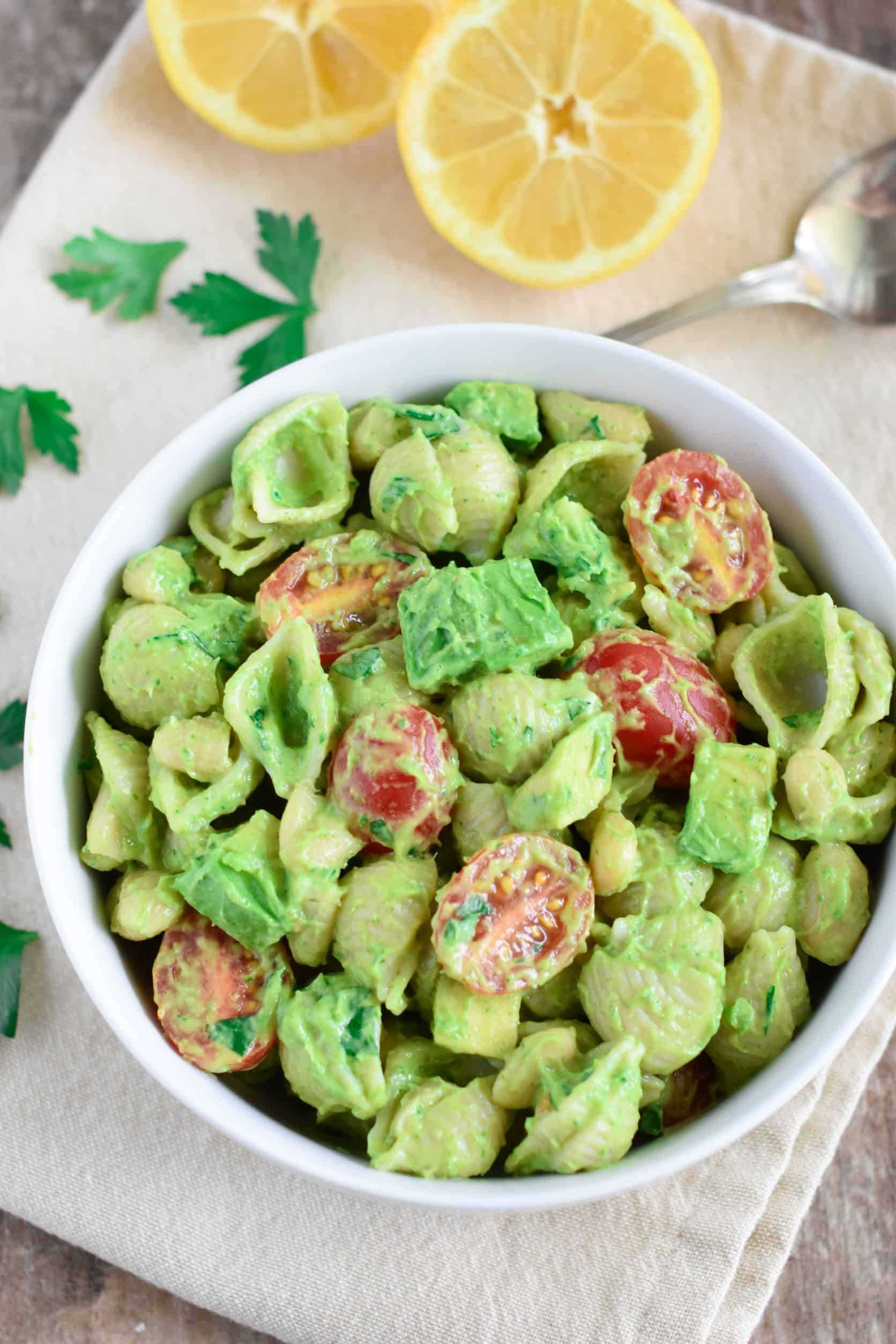 overhead of pasta salad in white bowl on kitchen napkin with a spoon, parsley leaves and lemon on it.