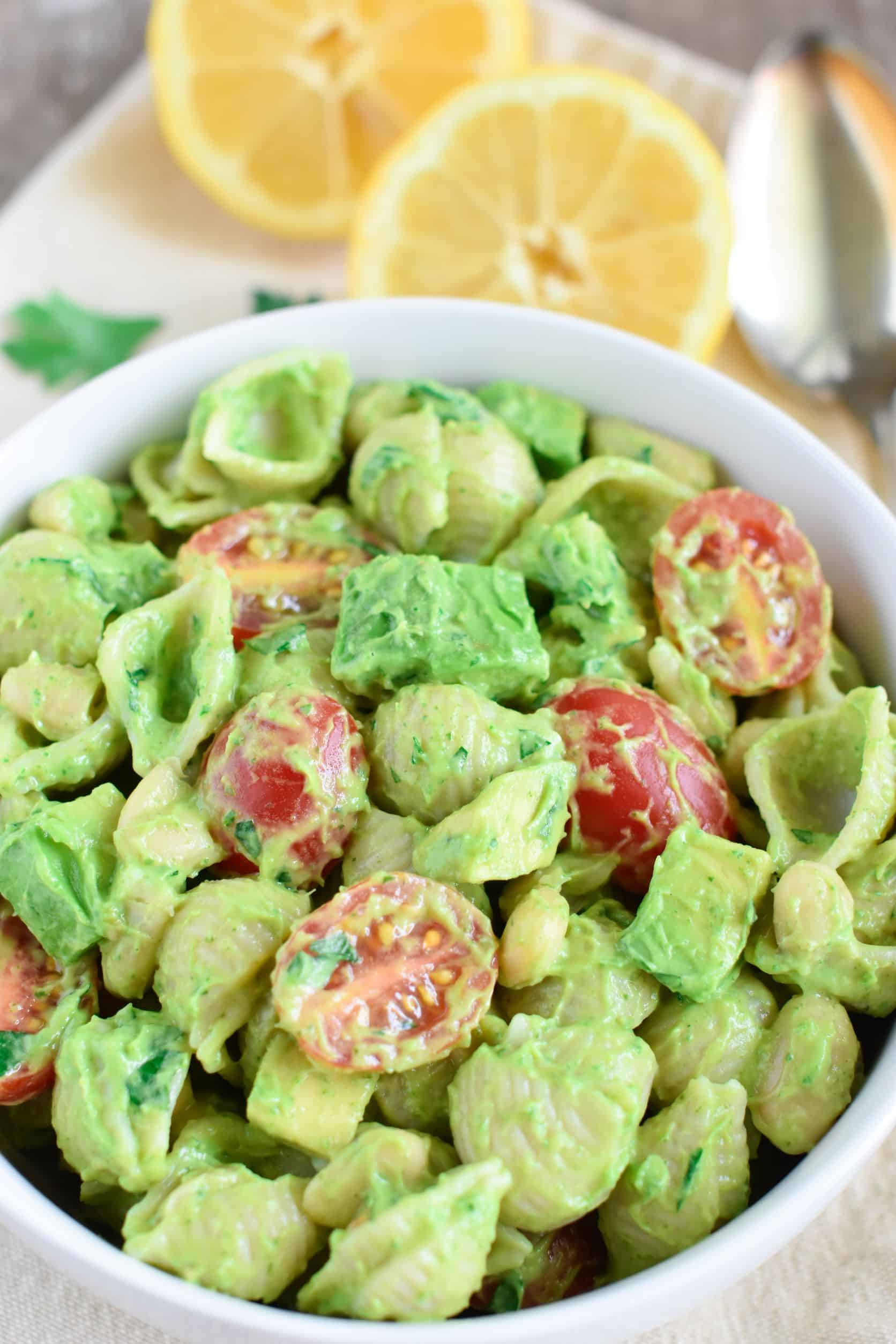 close up of pasta salad in white bowl with parsley, lemon and spoon in background.