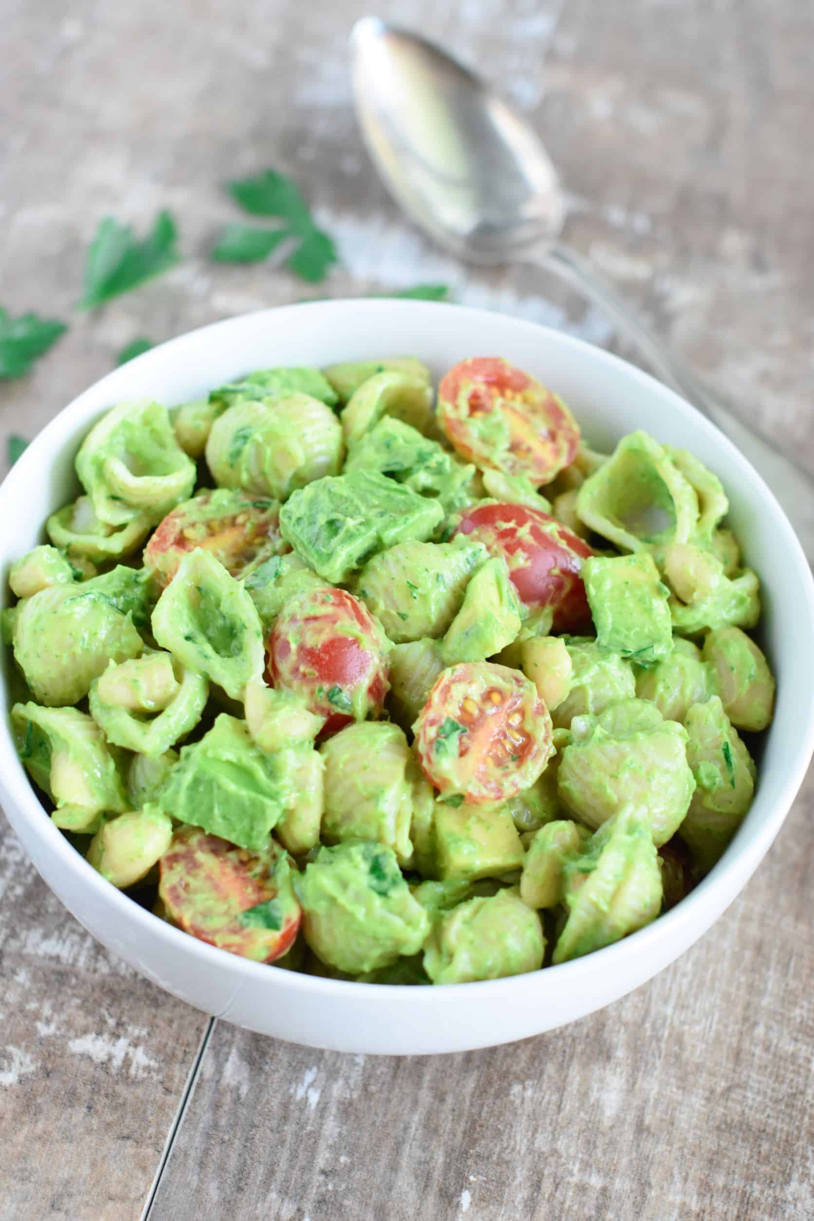 side view of pasta salad in a white bowl with spoon and parsley leaves behind it.