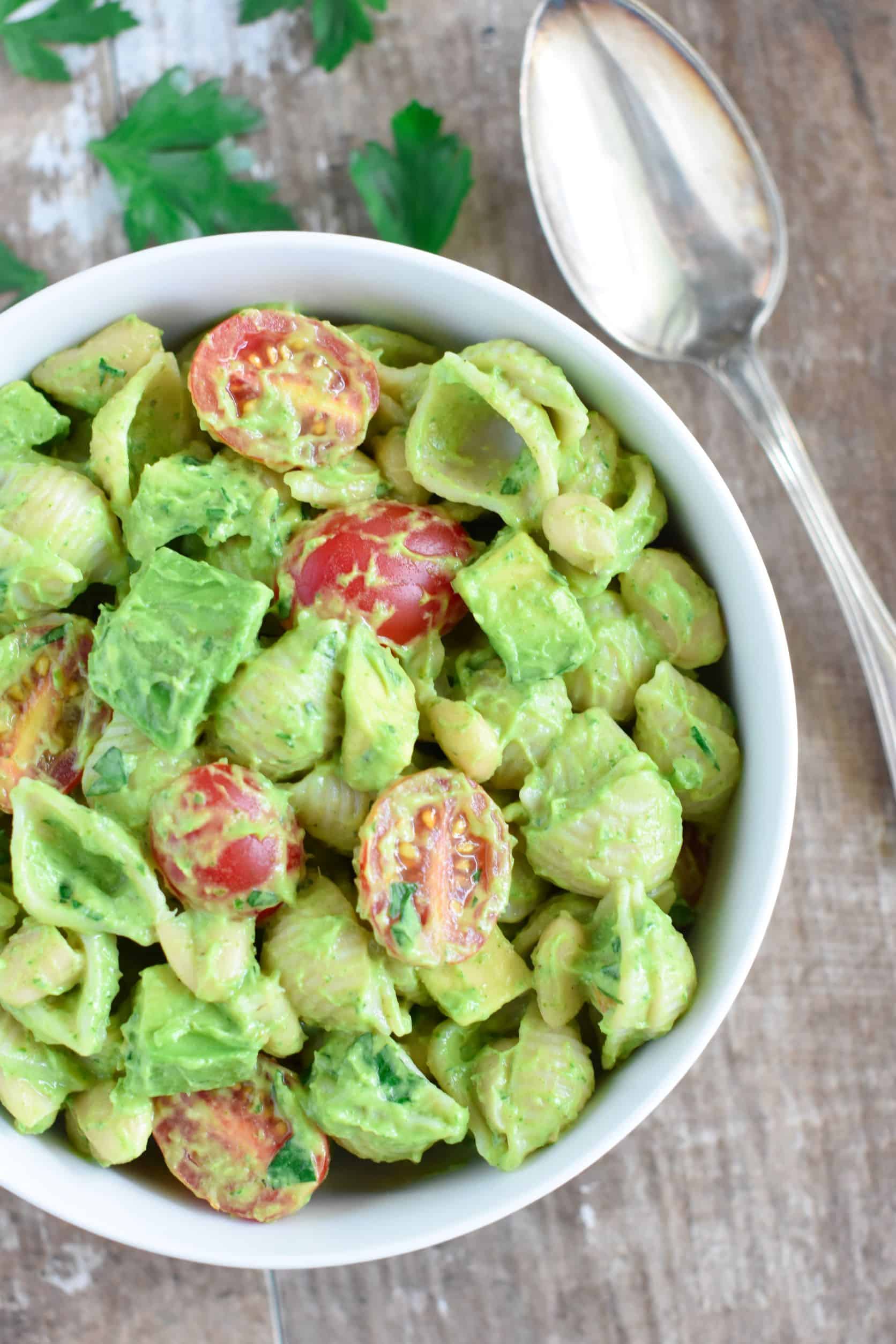 overhead close up of avocado pasta salad in white bowl with a spoon next to it and parsley leaves behind it.