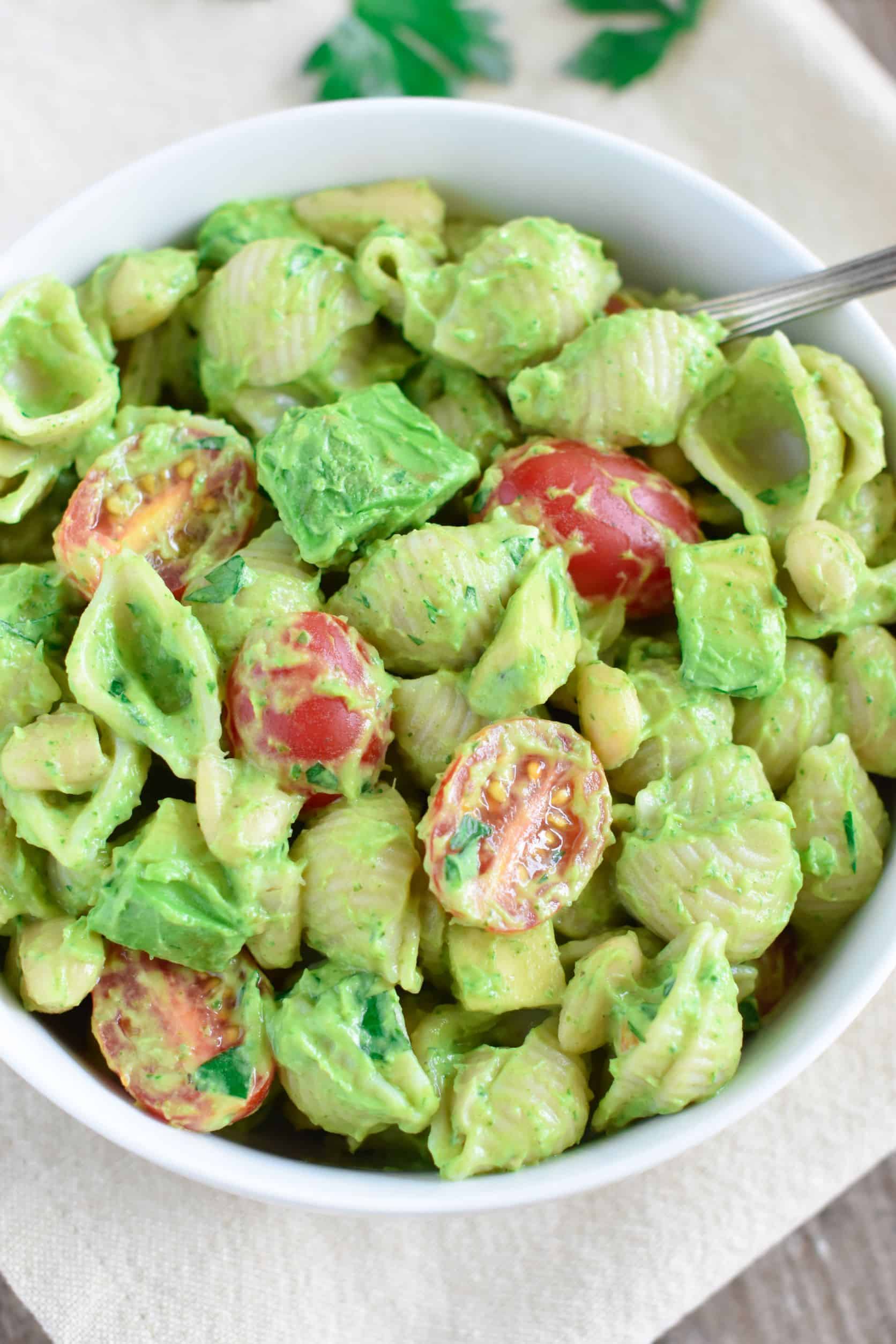 pasta salad in a white bowl with a spoon in it on a kitchen napkin with some parsley leaves on it.