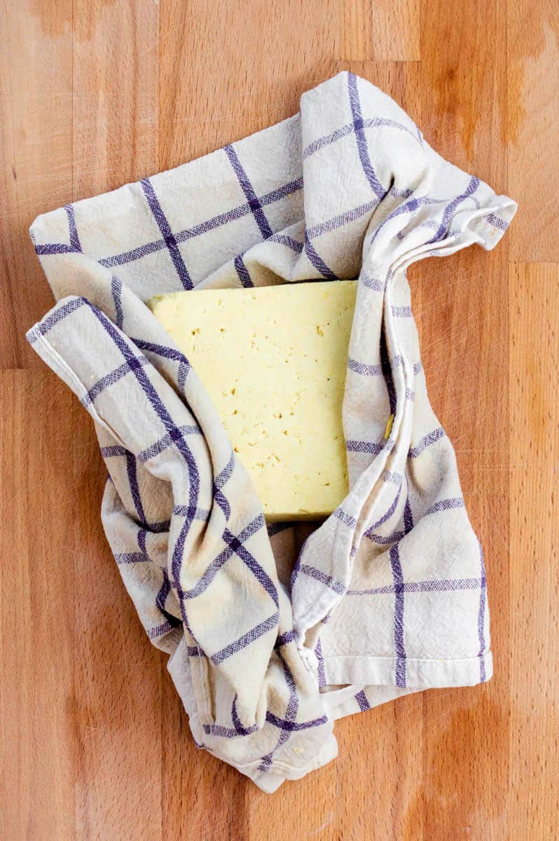 A block of tofu wrapped in a kitchen towel and placed on a wooden cutting board.