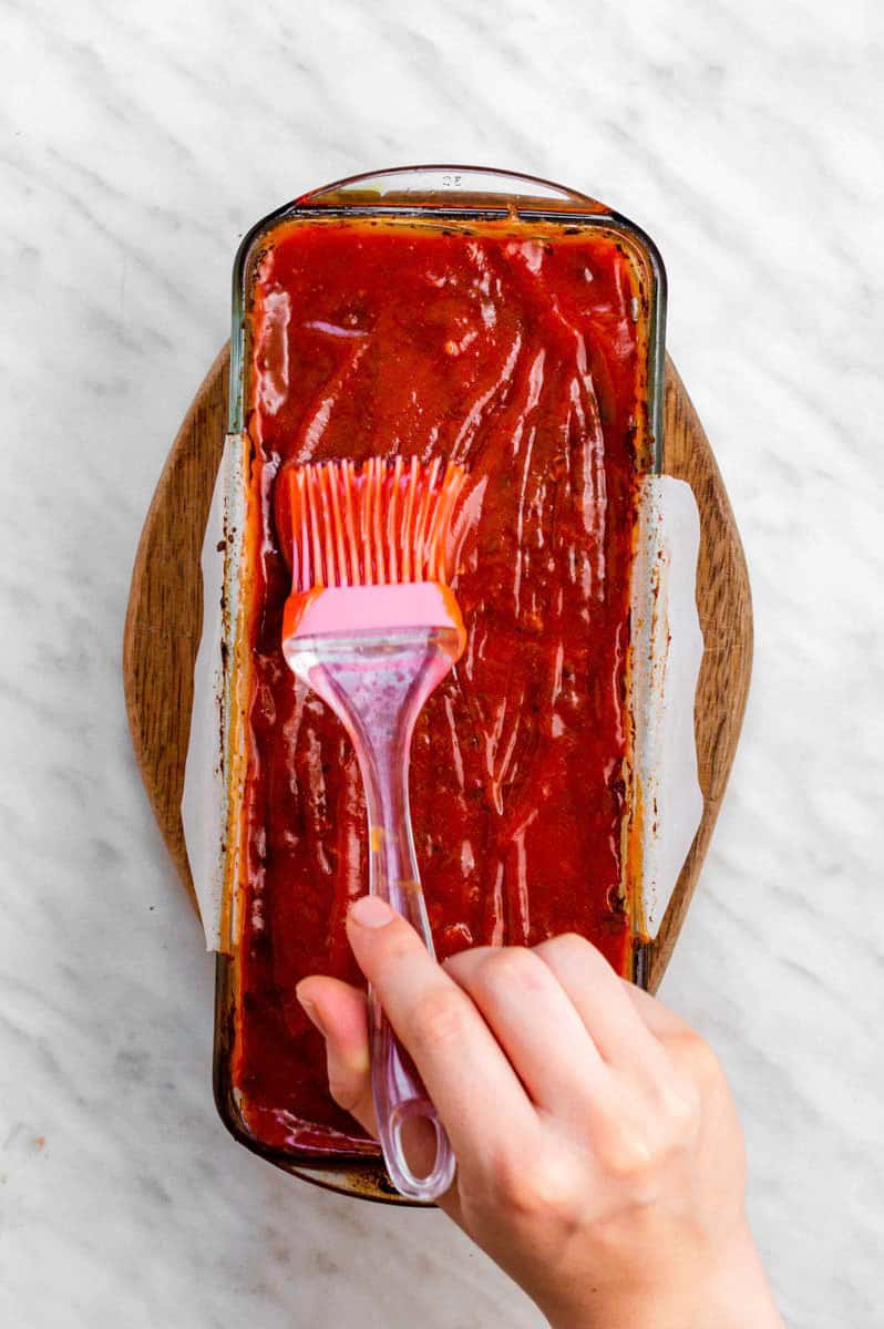 A hand brushing the ketchup glaze over Beyond meat meatloaf with a pink pastry brush.